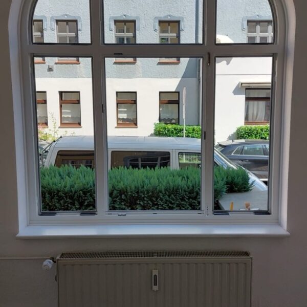 Großes, weißes Rundbogenfenster mit Sprossen, das auf eine Straße mit Gebäuden blickt.