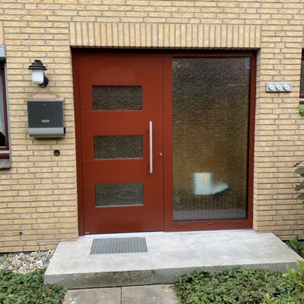 Rote Hauseingangstür mit drei Glaseinsätzen und großem Seitenteil aus Ornamentglas in Klinkerfassade.